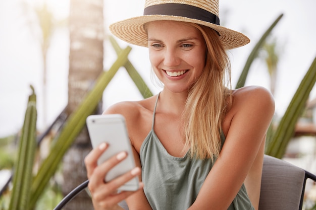 Adorable mujer rubia feliz vestida de manera informal, usa sombrero de paja y una camiseta informal, se alegra de leer buenos comentarios debajo de su foto o recibe un mensaje agradable de su novio mientras tiene vacaciones de verano