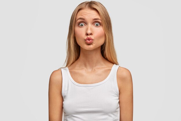 Adorable mujer rubia con piel sana y ojos abiertos, mantiene los labios redondos, hace muecas, vestida con una camiseta blanca informal, tiene una figura delgada, posa sobre una pared en blanco. Expresiones faciales, belleza