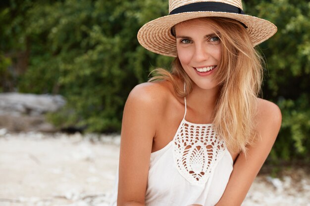 Adorable mujer rubia satisfecha vestida con ropa de verano, posa al aire libre en la playa contra la vegetación verde, disfruta de un clima soleado, pasa sus vacaciones en la playa. Gente, ocio, concepto de belleza