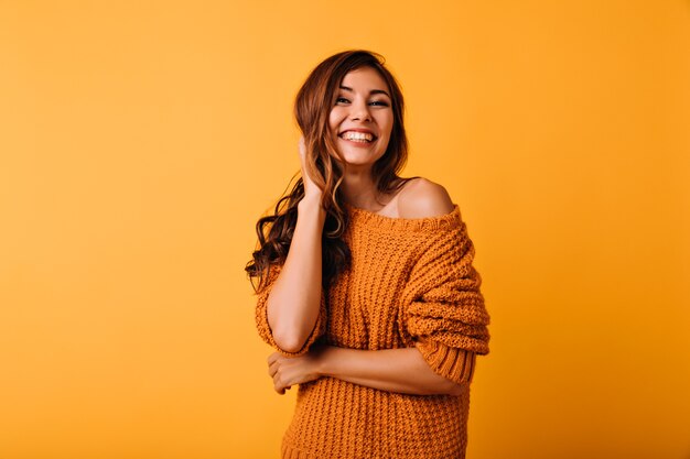 Adorable mujer vestida de naranja tocando su cabello castaño ondulado. Riendo alegre chica posando en amarillo.