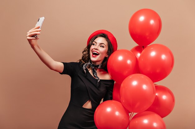 Adorable mujer vestida de negro y boina roja sostiene un teléfono inteligente, hace selfie y posa con globos.