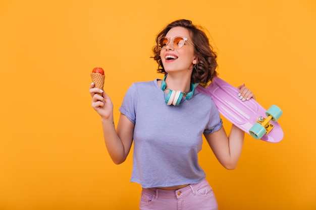 Adorable niña blanca con helado expresando felicidad. Retrato de interior de dama espectacular con monopatín morado lindo.