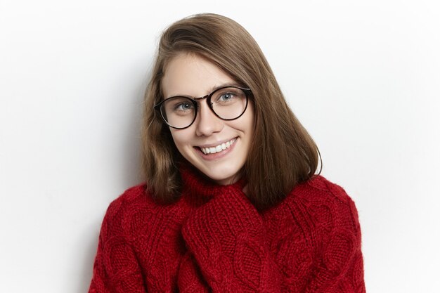 Adorable niña bonita con anteojos elegantes y suéter de punto cálido, sintiendo frío después de caminar, calentando las manos dentro de las mangas, sonriendo