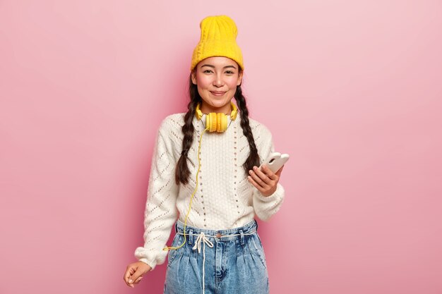 Adorable niña de cabello oscuro con coletas, usa un teléfono móvil para navegar por las redes sociales, usa un elegante sombrero