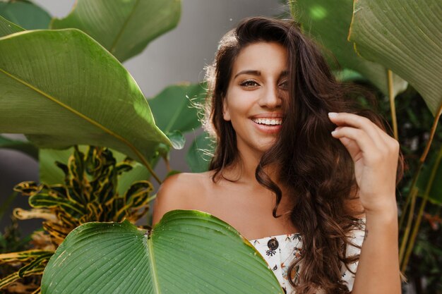 Adorable niña con cabello rizado se ríe contra el fondo de grandes hojas verdes Dama de ojos verdes en la parte superior blanca con estampado floral posando