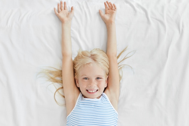 Adorable niña con cabello rubio y pecas, que se despierta por la mañana, se estira sobre la ropa de cama blanca y tiene una sonrisa agradable