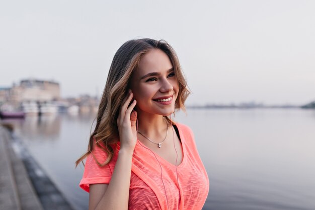 Adorable niña caucásica escuchando música con expresión de cara feliz mientras camina cerca del río. Retrato al aire libre de una mujer increíble pasar tiempo junto al lago.