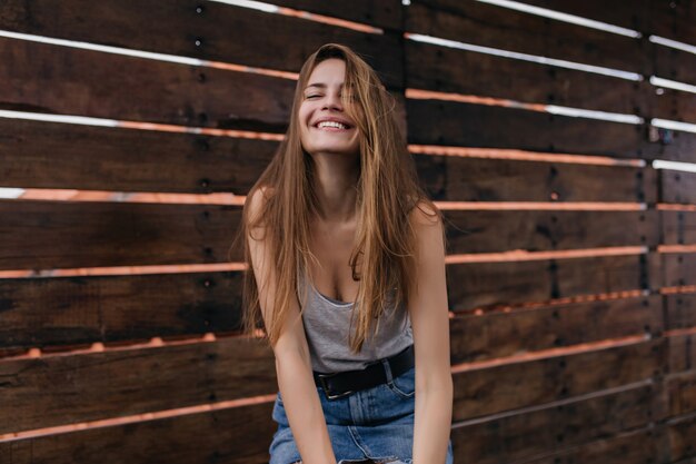 Adorable niña con cinturón negro posando junto a la pared de madera. Retrato de mujer joven complacida en falda de mezclilla riendo en un cálido día de primavera.