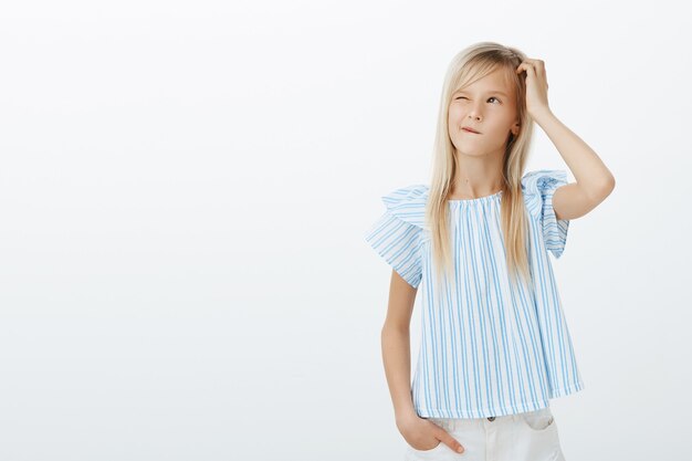 Adorable niña europea confundida con blusa azul, rascándose la cabeza y mirando hacia arriba con una leve sonrisa, confundida y cuestionada, pensando qué hacer para divertirse, preocupada por la pared gris