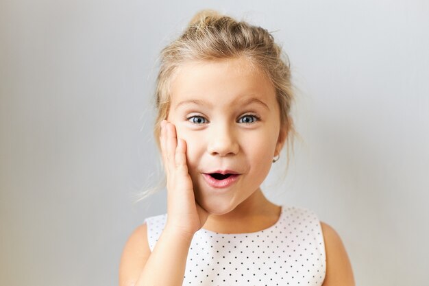 Adorable niña europea en edad preescolar posando aislada con la mano en la mejilla, diciendo Wow con la boca abierta, sorprendida con noticias emocionantes, expresando una verdadera reacción, vistiendo un vestido punteado