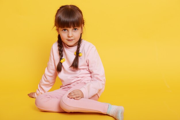 Adorable niña europea sentada en el piso, vestida con pantalones y suéter rosa pálido, aislado sobre fondo amarillo, mira a la cámara, niña de cabello oscuro con coletas.