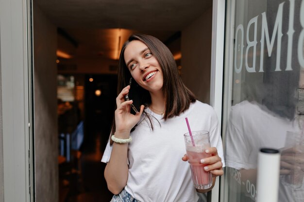 Adorable niña feliz con una sonrisa maravillosa está hablando por teléfono y bebiendo un batido de verano brillante mientras sale de la cafetería