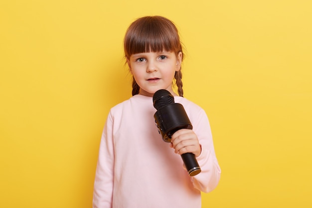 Adorable niña con micrófono sobre fondo amarillo, mira a la cámara mientras habla en micrófono, apuntando con el dedo índice a un lado. Copie el ritmo del texto publicitario o promocional.