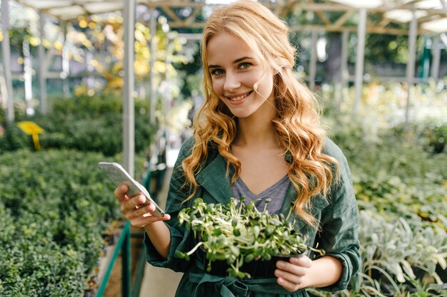 Adorable niña de ojos verdes brillantes y largos rizos de buen humor. Retrato en invernadero de modelo bonita sosteniendo teléfono y planta en sus manos