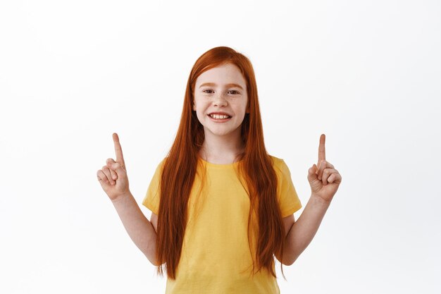 Adorable niña pelirroja, niño con cabello largo pelirrojo apuntando con el dedo hacia arriba y sonriendo complacido, muestra algo, texto promocional en el espacio de la copia, fondo blanco.