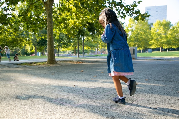 Adorable niña de pelo negro jugando a la rayuela en el parque de la ciudad. Longitud total, espacio de copia. Concepto de infancia
