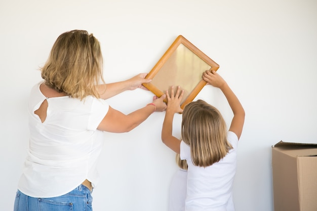 Adorable niña poniendo marco de fotos en la pared blanca con la ayuda de mamá