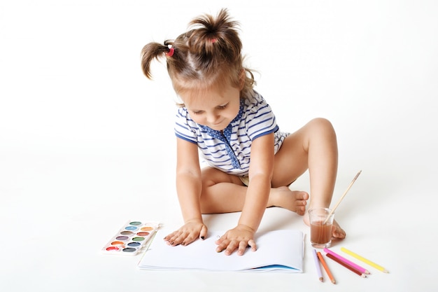 Adorable niña de preescolar con manos pintadas, hace huellas digitales en la página en blanco del álbum, usa acuarela para hacer fotos, es muy creativa, está aislada sobre la pared blanca del estudio