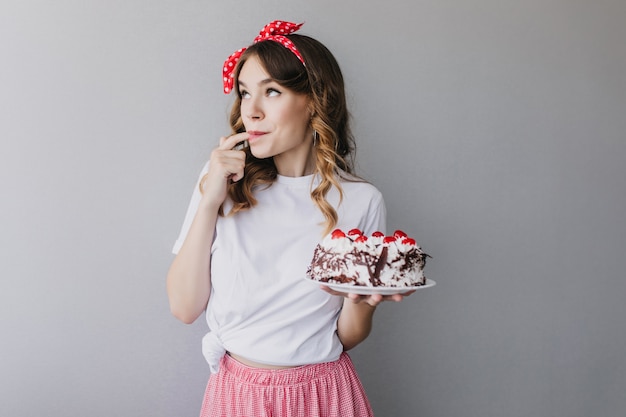 Adorable niña rizada probando tarta de fresa. Filmación en interiores de modelo femenino romántico con cinta roja en el pelo sosteniendo un sabroso pastel.