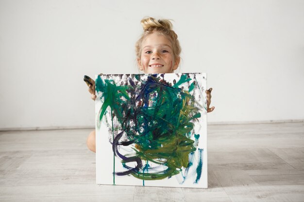 Adorable niña rubia sonriente hading por la imagen. Linda niña con moño y pecas posando contra la pared blanca. La sonrisa infantil es una fuente de emociones positivas.