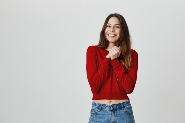 Adorable niña se siente feliz o agradecida, junta las manos y sonríe optimista