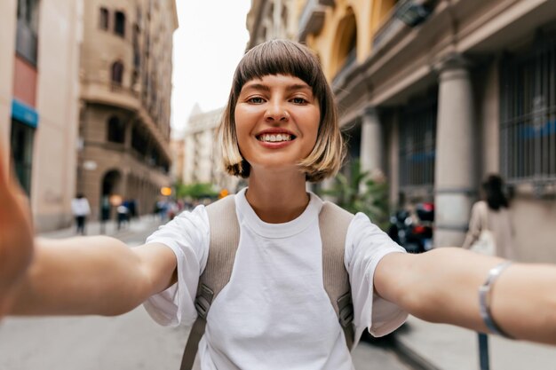 Adorable niña sonriente feliz con el pelo corto con camiseta blanca está haciendo selfie y disfrutando del verano caminando en la ciudad Señora alegre haciendo selfie en un día soleado