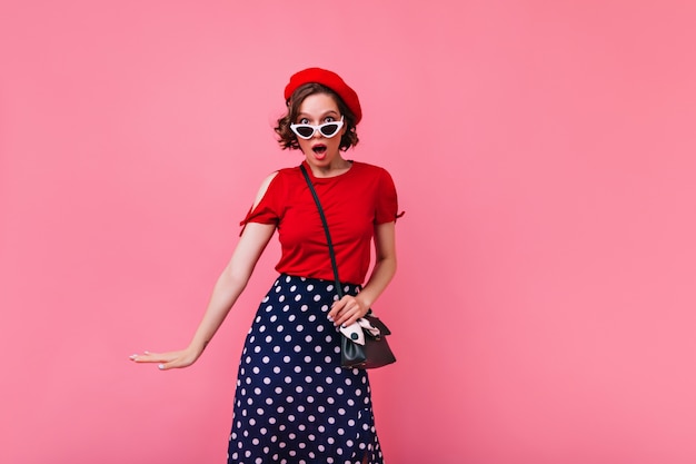 Adorable niña sorprendida en boina roja de pie en la pared rosada. Foto interior de dama francesa asombrada con peinado ondulado.