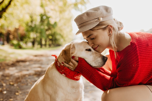 Adorable niña y su perro sentados bajo árboles verdes y sol brillante en el parque. Preciosa rubia pasando un buen rato junto a su mascota.