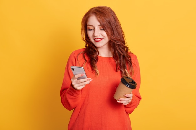 Adorable niña con suéter naranja posando aislado, sosteniendo café para llevar y teléfono móvil