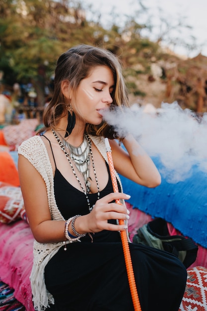 Adorable niña vestida de negro y elegantes aretes con placer fuma una pipa de agua, sentada en un sofá colorido.