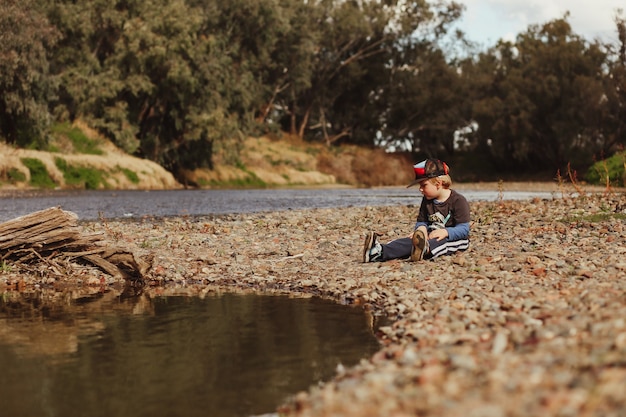 Adorable niño australiano rubio sentado sobre guijarros en la orilla del río