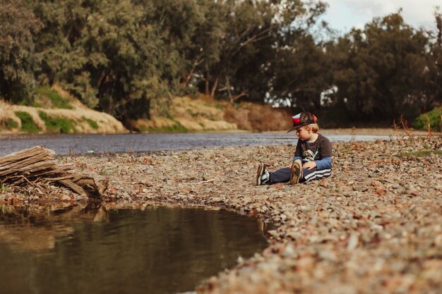 Adorable niño australiano rubio sentado sobre guijarros en la orilla del río