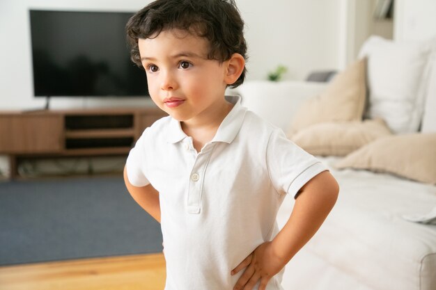 Adorable niño con camisa blanca de pie con las manos en las caderas en la sala de estar
