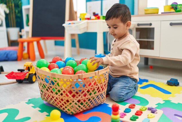 Adorable niño hispano jugando con pelotas sentado en el piso en el jardín de infantes