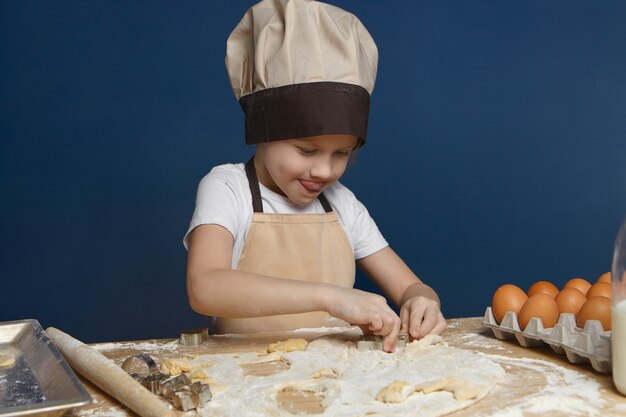 Adorable niño varón de 8 años en delantal beige y sombrero de pie en la cocina