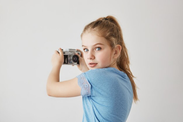 Adorable pequeña niña rubia con ojos azules va a tomar selfie. Ella mira hacia atrás con expresión asustada cuando escucha a mamá entrar en la habitación