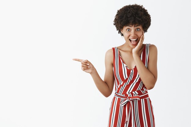 Adorable sorprendida e impresionada atractiva mujer afroamericana con elegantes monos a rayas formales tocando la mejilla de la sorpresa y apuntando a la izquierda con el dedo índice sonriendo con asombro