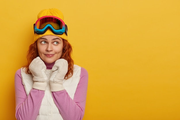 La adorable turista tiene el pelo rojo, lleva un sombrero amarillo con gafas de esquí en la cabeza, guantes blancos suaves, un jersey morado con chaleco blanco, tiene un descanso activo durante la nieve, mira hacia otro lado, posa en el interior