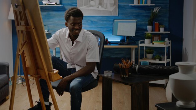 Adulto afroamericano sentado en un estudio de arte dibujando con lápiz, diseño de jarrón y lienzo blanco sobre caballete. Joven negro con pasatiempo creativo haciendo obras maestras exitosas y bellas artes