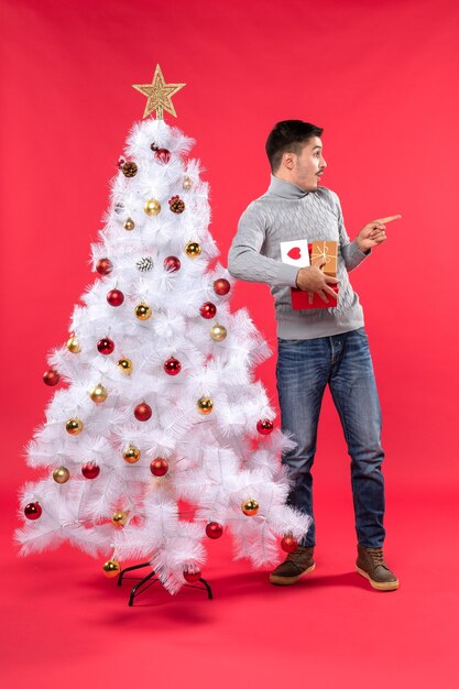 Adulto guapo romántico de pie cerca del árbol de Navidad blanco decorado y sosteniendo sus regalos mirando sorprendido en rojo