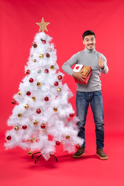 Adulto guapo romántico de pie cerca del árbol de Navidad blanco decorado y sosteniendo sus regalos en rojo