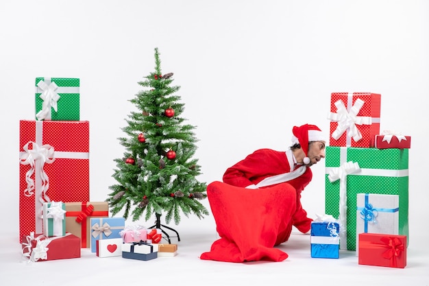 Adulto joven disfrazado de Papá Noel con regalos y árbol de Navidad decorado sentado en el suelo buscando algo