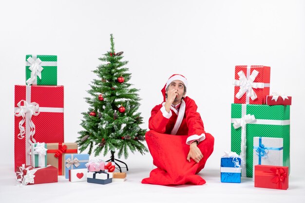 Adulto joven emocionado confundido vestido como Papá Noel con regalos y árbol de Navidad decorado sentado en el suelo sobre fondo blanco.