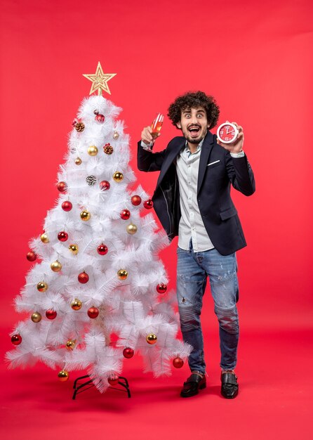 Adulto joven emocionado sosteniendo el reloj y una copa de vino y de pie cerca del árbol de Navidad en la imagen roja