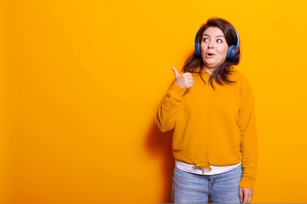 Adulto joven escuchando música en auriculares y haciendo bien el gesto con la mano en el estudio. Mujer positiva que se siente alegre y escucha la canción y el audio en el auricular, divirtiéndose en la cámara.