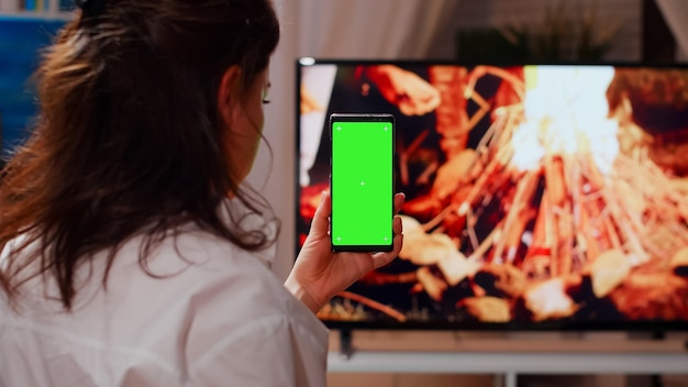 Adulto joven mirando el teléfono inteligente con pantalla verde y comiendo papas fritas en casa. Dispositivo de sujeción de mujer con fondo aislado, plantilla de maqueta y clave de croma frente a la televisión