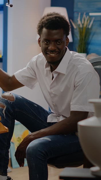 Adulto joven negro sentado en jarrón de dibujo de estudio de arte con lápiz y lienzo blanco sobre caballete. Hombre afroamericano con ocupación creativa haciendo obras maestras innovadoras y bellas artes