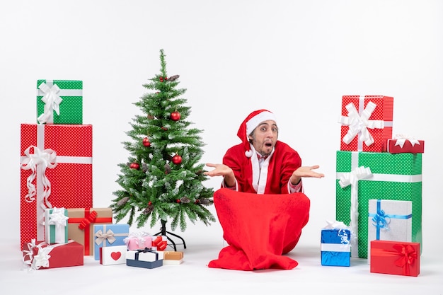 Adulto joven sorprendido disfrazado de Papá Noel con regalos y árbol de Navidad decorado sentado en el suelo sobre fondo blanco.