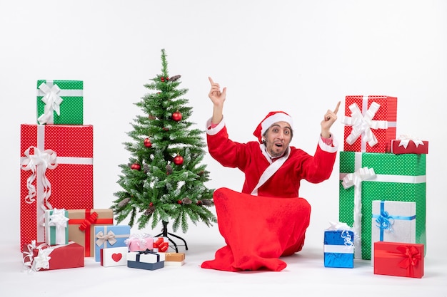 Adulto joven sorprendido vestido como Papá Noel con regalos y árbol de Navidad decorado sentado en el suelo apuntando algo en ambos lados sobre fondo blanco.