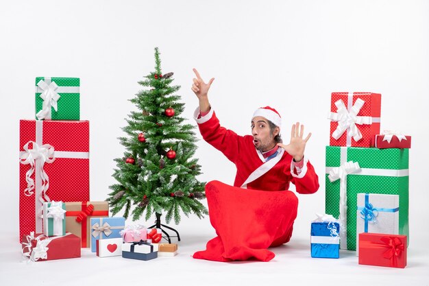 Adulto joven vestido como Santa Claus con regalos y árbol de Navidad decorado sentado en el suelo apuntando arriba mostrando cinco